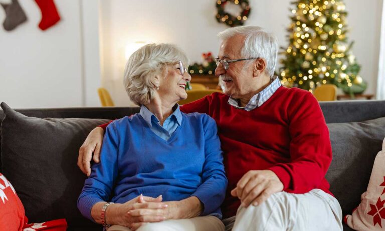Senior couple sitting together on a couch, smiling warmly at each other in a cozy living room decorated for the holidays with a Christmas tree