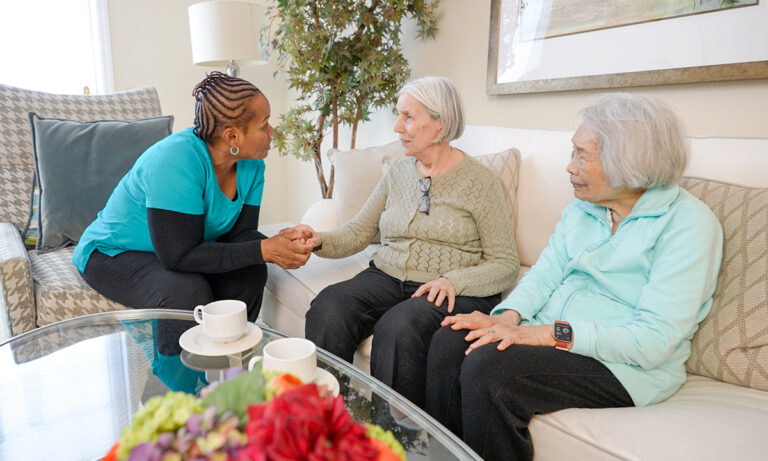 Professional caregiver providing companionship and support to two older women seated together in a calm, welcoming home setting