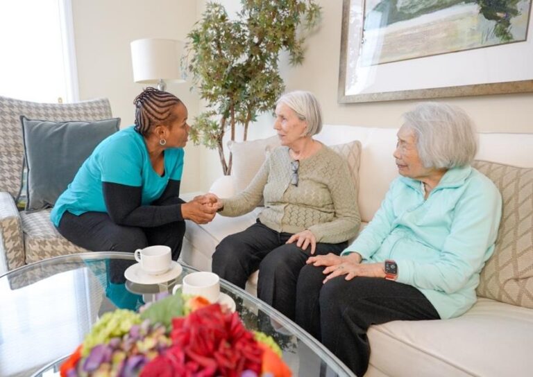 Professional caregiver providing companionship and support to two older women seated together in a calm, welcoming home setting.