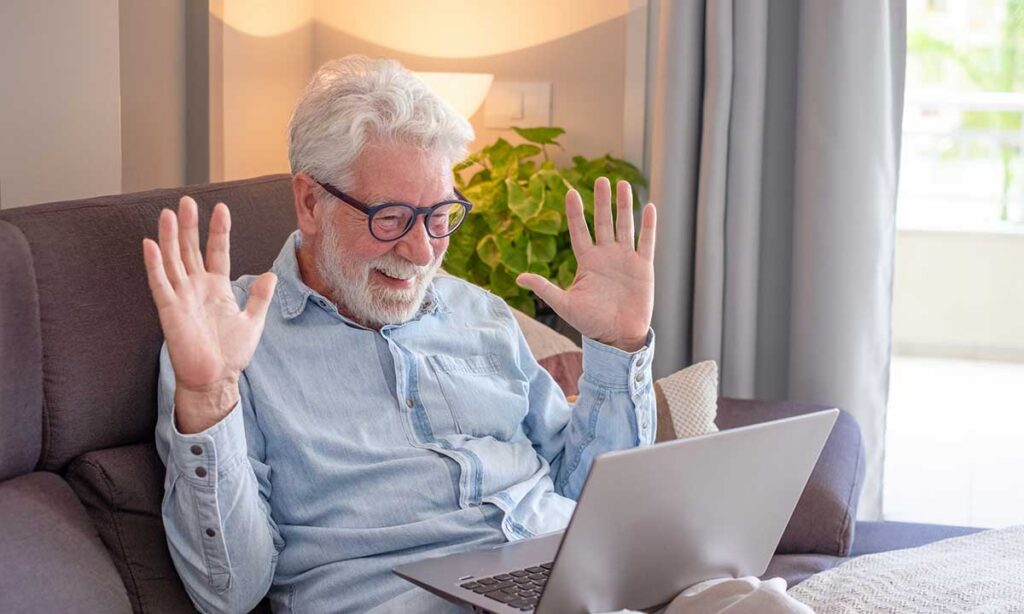 Senior man happily greeting family on a laptop video call in his living room