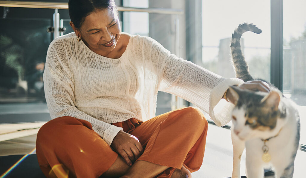 Senior woman sitting on the floor at home, smiling and petting a cat in natural sunlight, showing companionship and comfort while aging in place